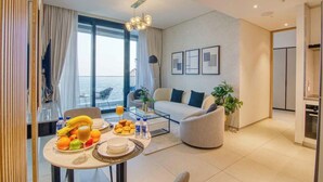 Een moderne woonkamer met een bank, een eettafel met fruit en glazen, en een groot raam met gordijnen.