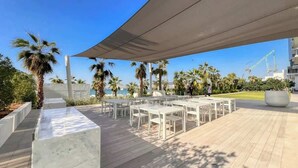 Un espace extérieur aménagé avec des tables et des chaises blanches, un auvent ombragé et une vue sur la mer.
