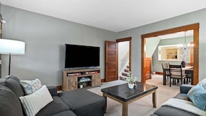 A living room with a sofa, a coffee table, and a TV on a wooden cabinet. There is a staircase leading to a dining area. - Entire Duplex - Historic District - Sleeps 12 (Bedford)
