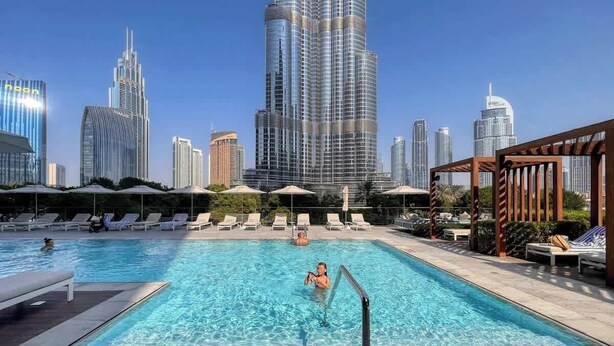 Una piscina en la azotea con sillones y sombrillas, rodeada de edificios altos.