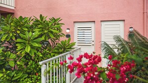 Um edifício cor-de-rosa com porta e janela brancas, rodeado por folhagem verde e flores cor-de-rosa.