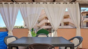 A covered outdoor seating area with a table and chairs, a view of a building with balconies, and a blue tarp on the left. - Raggio di sole Olbia (Olbia)