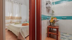 A hotel room with a bed, a wooden stool, a vase with flowers, and a bathroom with a tiled wall. - Raggio di sole Olbia (Olbia)