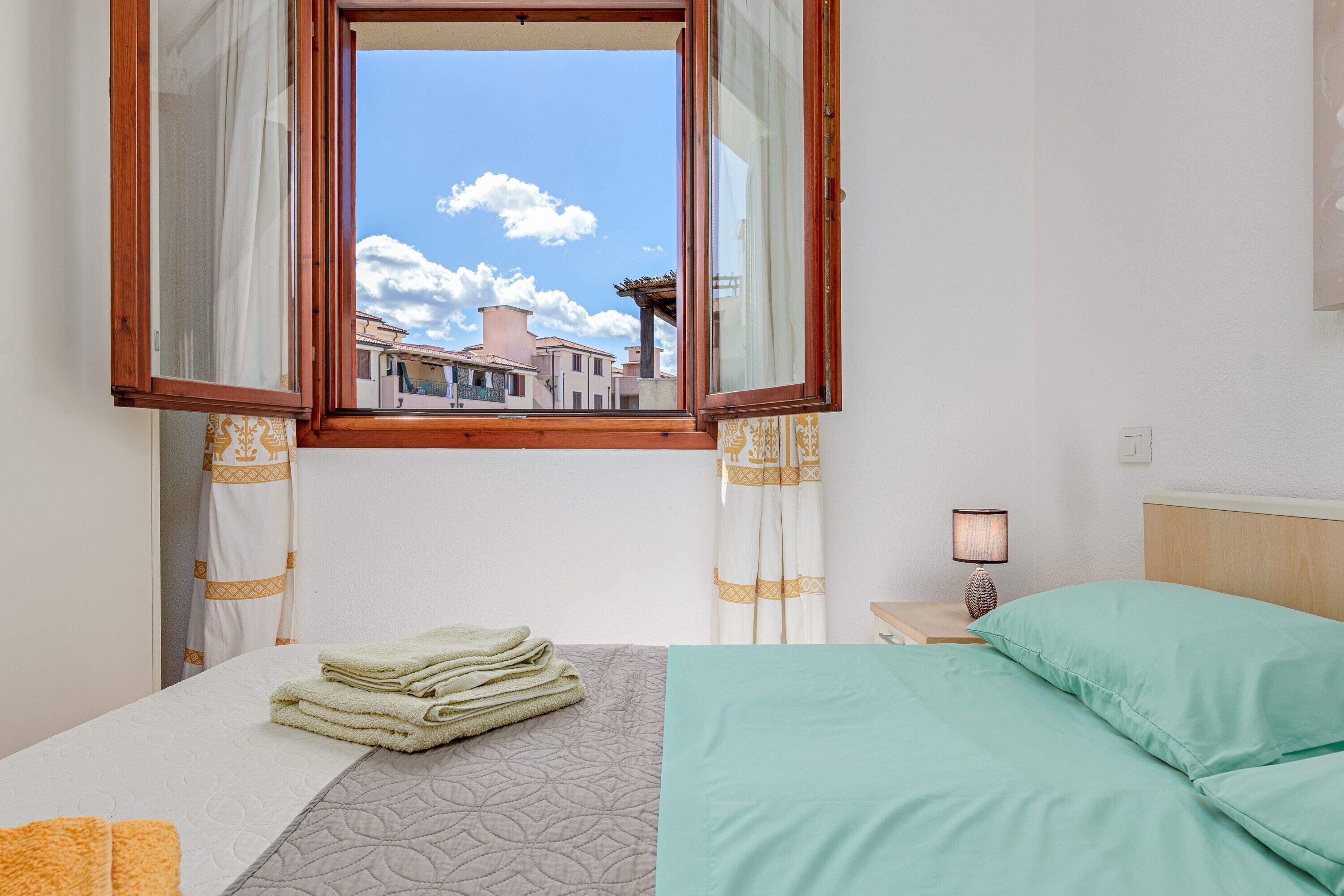 Ein Hotelzimmer mit Bett, Fenster mit Vorhängen, Nachttisch mit Lampe und Blick auf Gebäude.