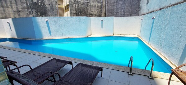 Una piscina con diseño de esquina, rodeada de suelo de baldosas y sillones de mimbre negro.