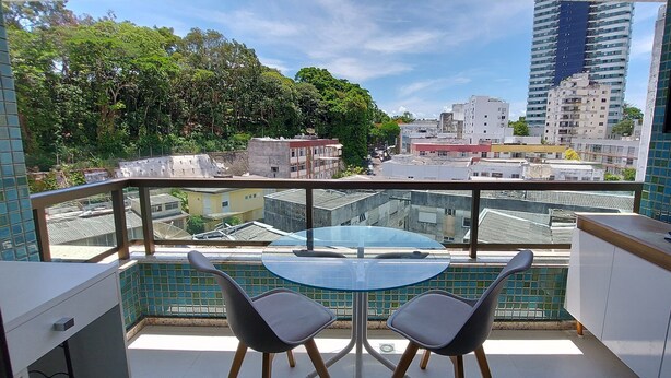 A balcony with a glass table and two chairs, overlooking a cityscape with buildings and trees. - COBAR0117 - Edifício Yacht Residencial (Salvador)