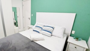 A neatly made bed with striped pillows, a grey blanket, and a white headboard in a room with teal walls and white furniture. - COBAR0117 - Edifício Yacht Residencial (Salvador)