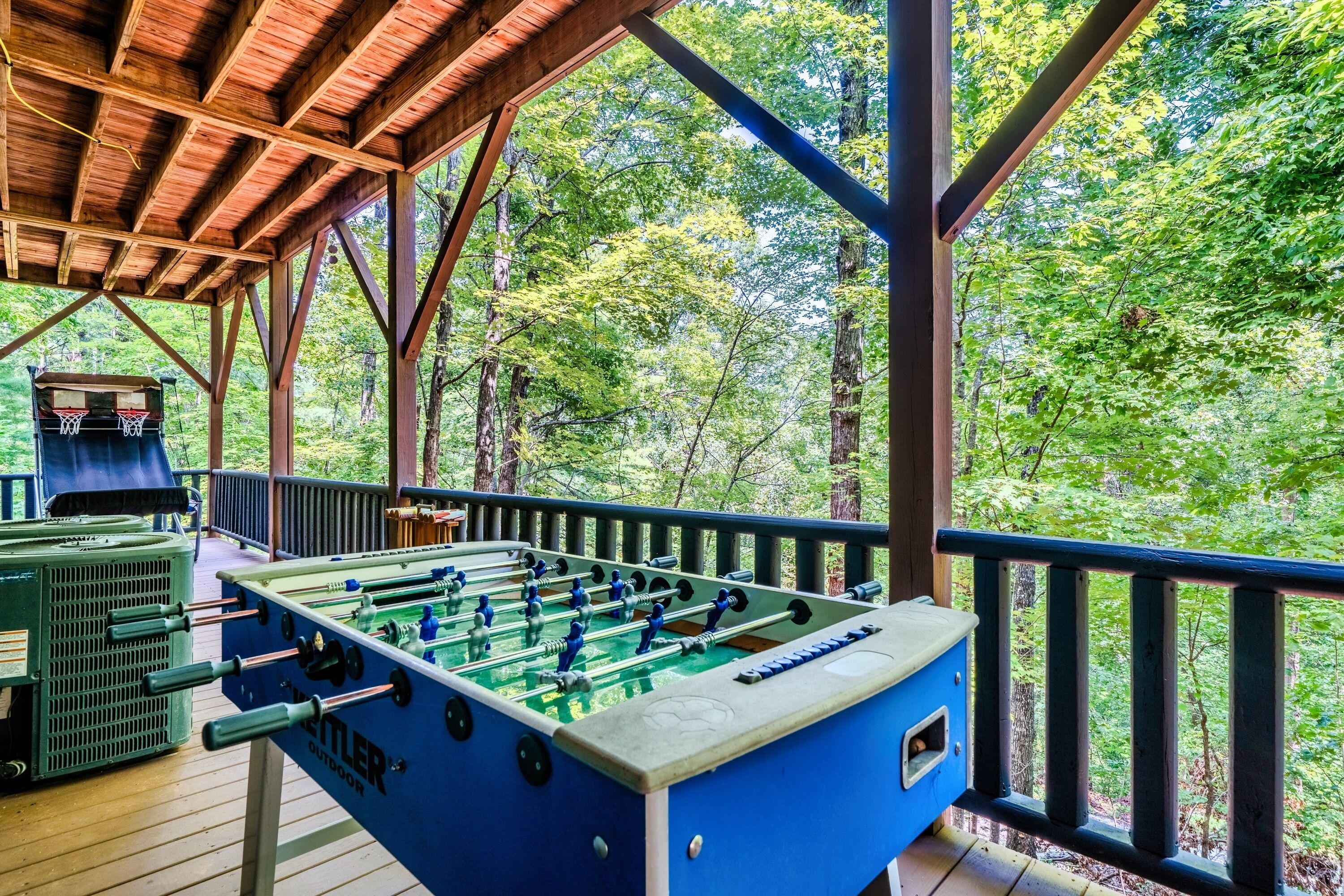 An outdoor foosball table under a wooden deck with a view of a forest.