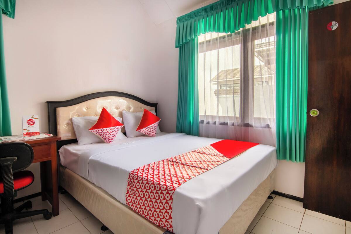 A neatly made bed with a patterned bedspread, a desk with a chair, and a window with curtains.