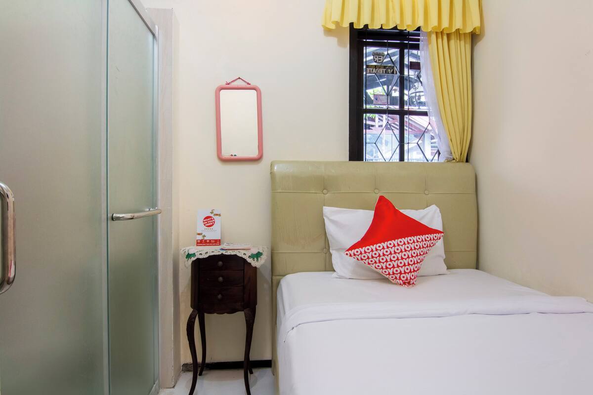 A hotel room with a bed, a bedside table with a lamp, a window with yellow curtains, and a door.