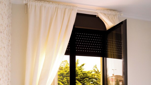 Una habitación con una silla de mimbre, una cama y un ventanal corredero que da a un balcón con mesa y sillas.
