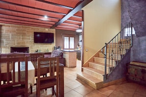 Amplia sala de estar con chimenea, una mesa de comedor con sillas, zona de cocina y una escalera con barandilla de metal.