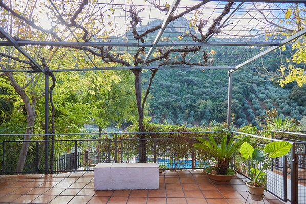 Un balcón techado con vistas a un área boscosa, un banco blanco y plantas en macetas.