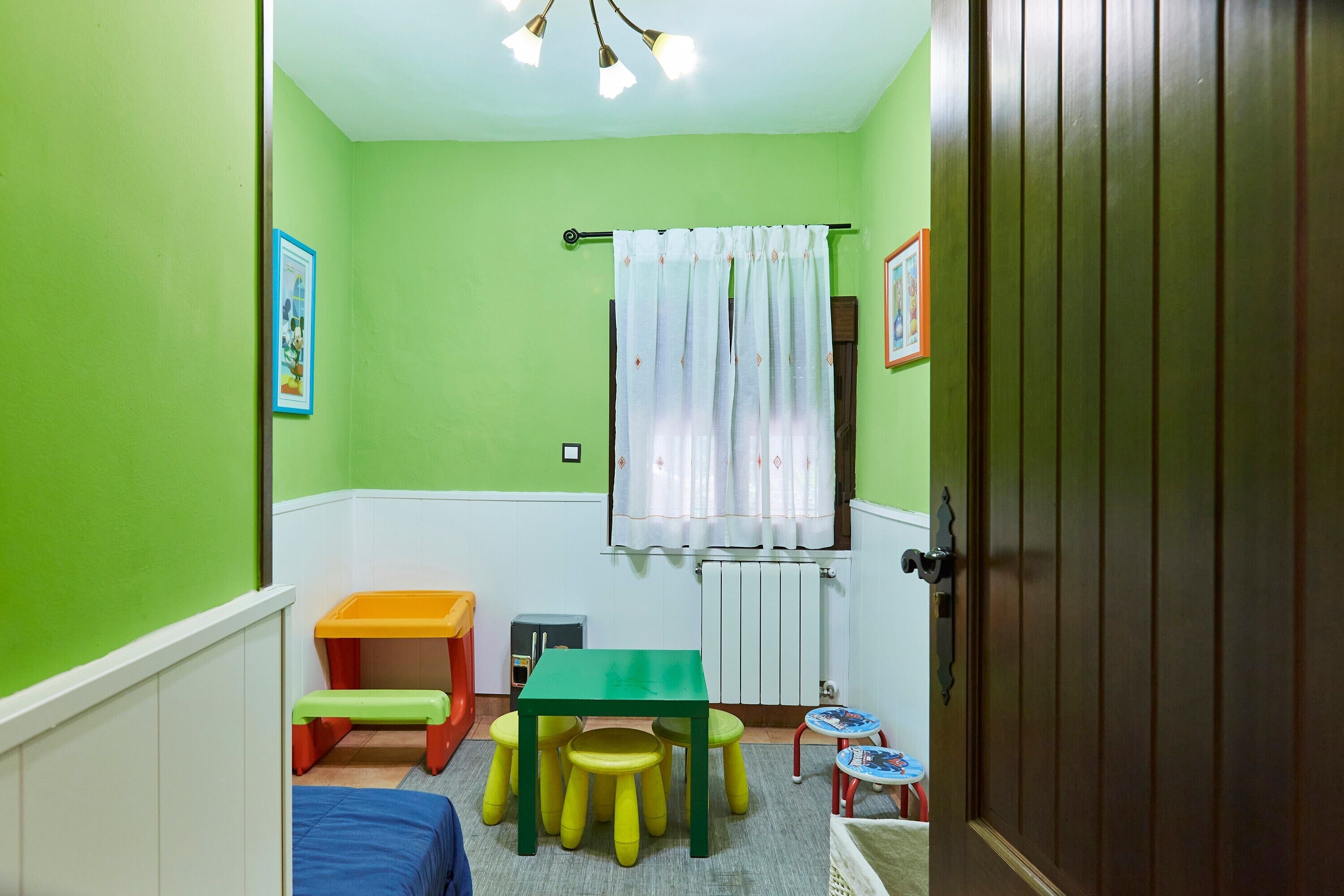 Una habitación infantil con pared verde, una mesa con taburetes, una cama, un radiador y una puerta.
