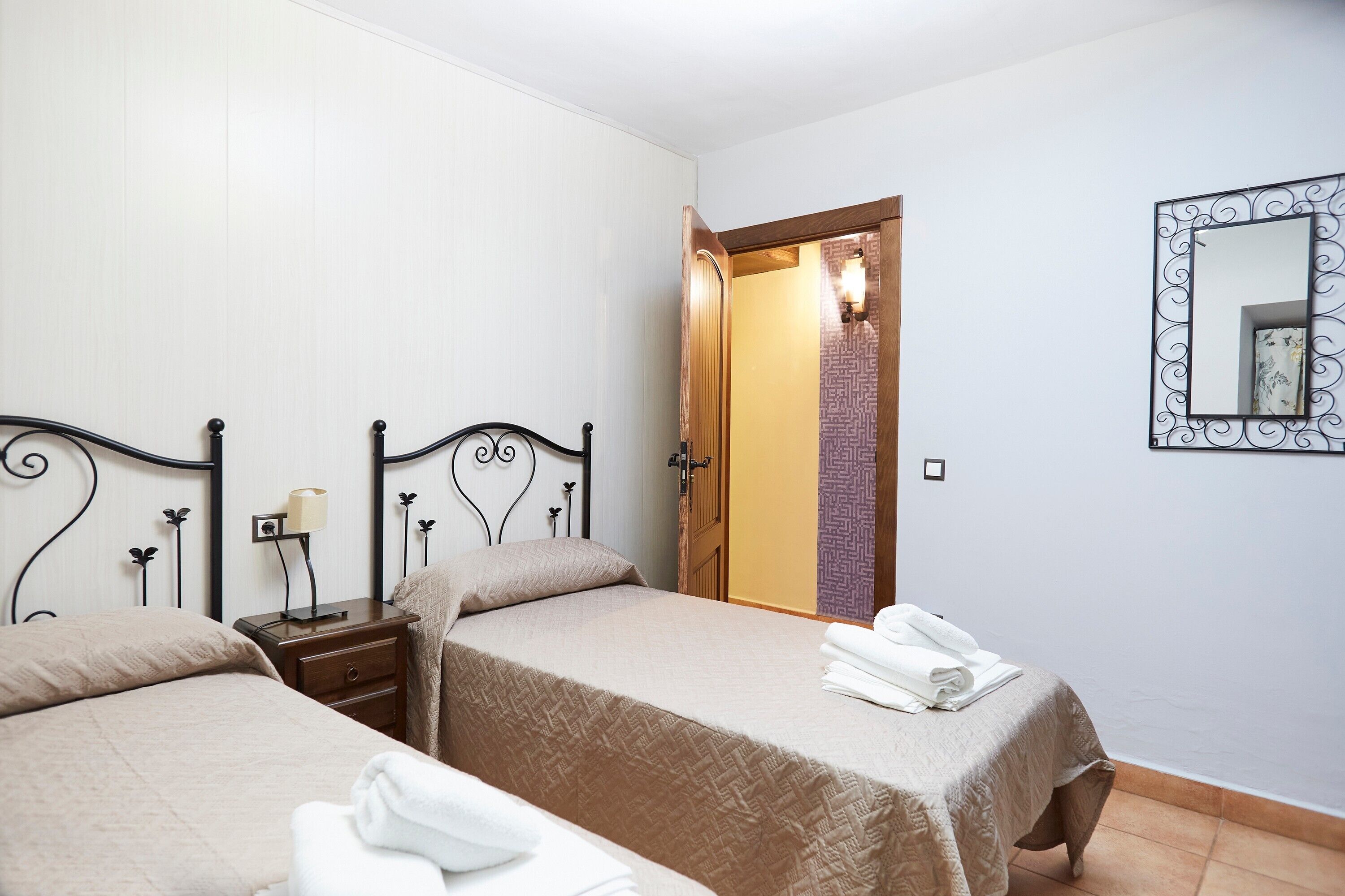 Habitación de hotel con dos camas, una mesita de noche con lámpara, un espejo y una puerta que da a otra habitación.