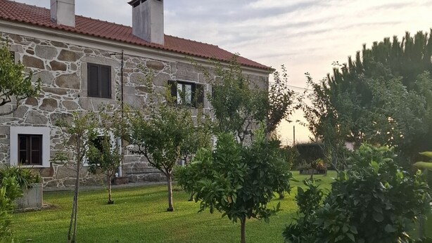 Un bâtiment en pierre avec un toit recouvert de tuiles rouges, une cheminée et un petit jardin planté de jeunes arbres.