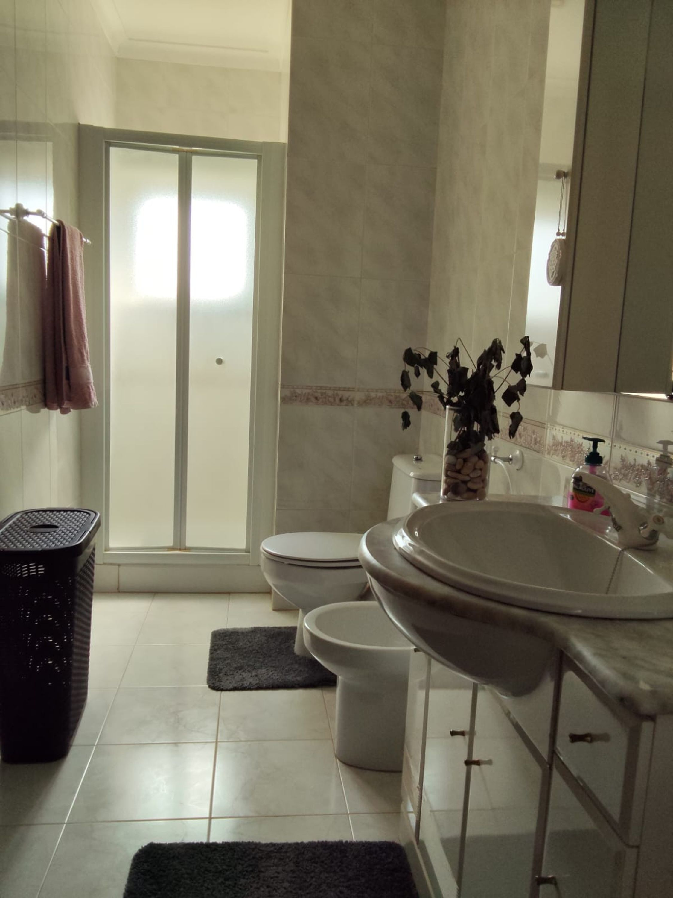A bathroom with a toilet, bidet, and a large sink. There is a shower area with a glass door, and a trash bin is visible.