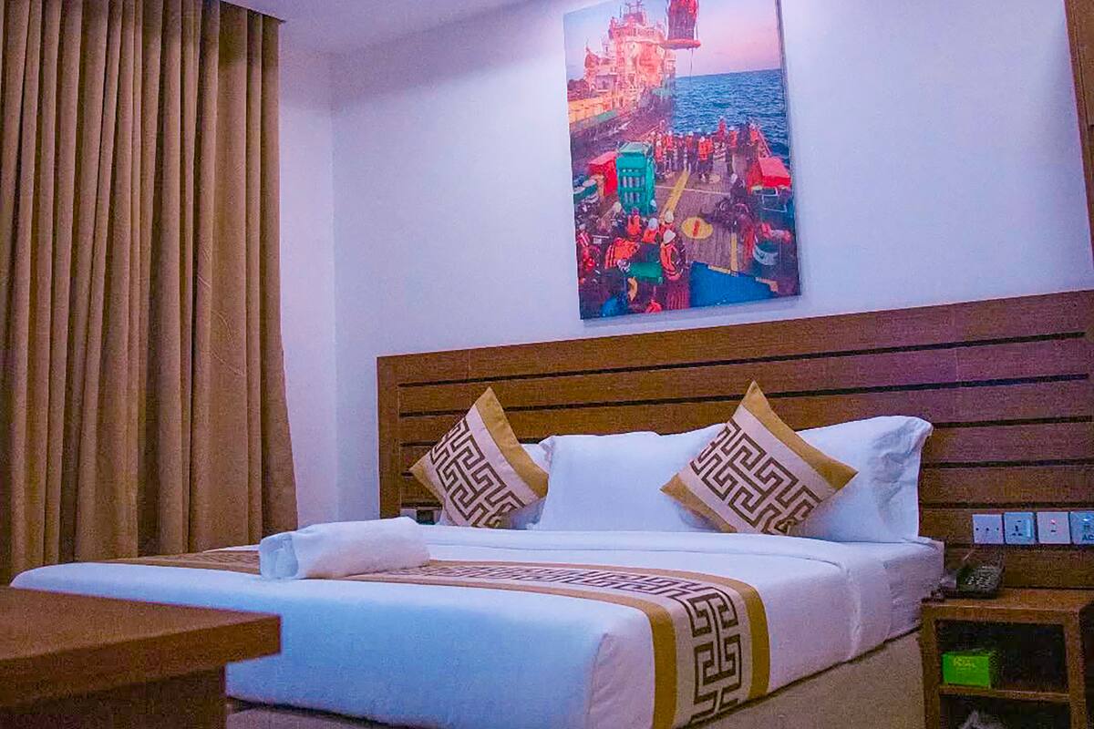 A hotel room with a bed, a nightstand, a painting of a coastal scene, and a window with curtains.