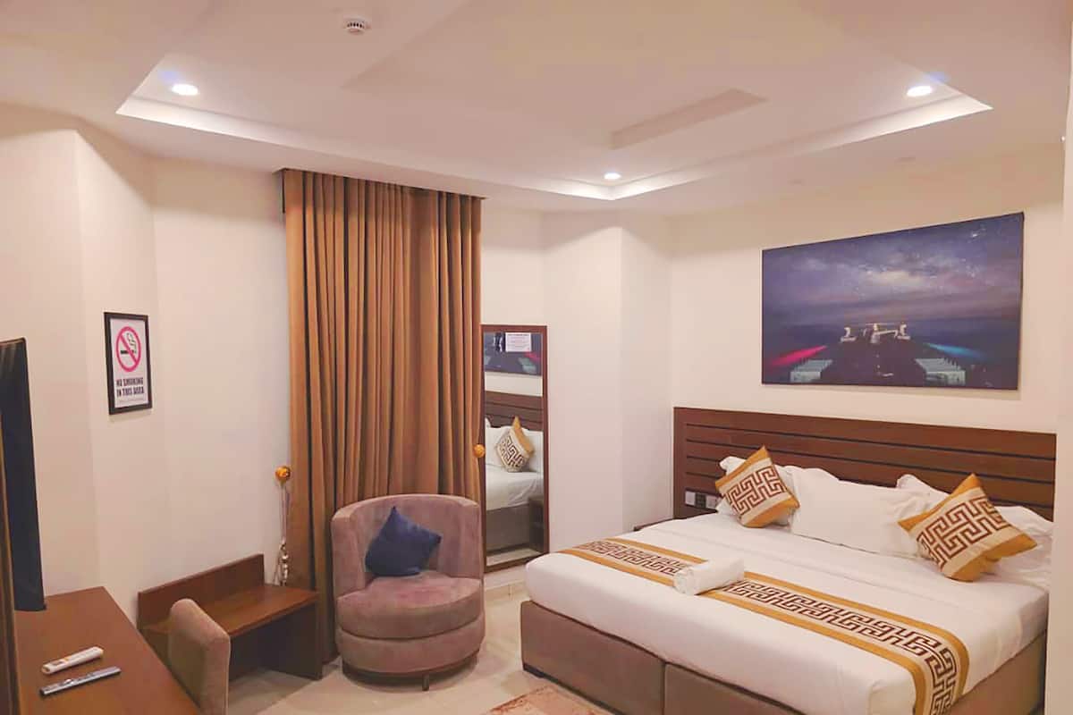 A hotel room with a bed, a desk, a chair, a large window with curtains, and a painting on the wall.