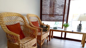 Deux fauteuils en osier avec des coussins, une petite table en bois ornée d’un vase et d’une plante, et une fenêtre avec des stores.