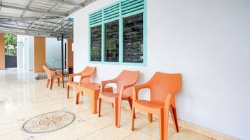 Un espacio de estar al aire libre cubierto con sillas de mimbre y una mesa, una ventana con contraventanas azules y suelo de baldosas.