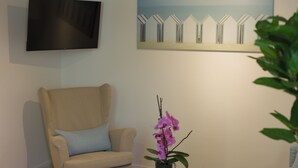A living room with a beige armchair, a small round table with a purple orchid, a flat-screen TV, and a painting of beach huts on the wall. - Moorrose: Holiday apartment in the Frisian Gulfhof, not barrier-free (Apen)
