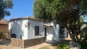 Un edifici petit, de color blanc, amb teulada de rajola, base de pedra i una zona de jardí amb un camí i un arbre.