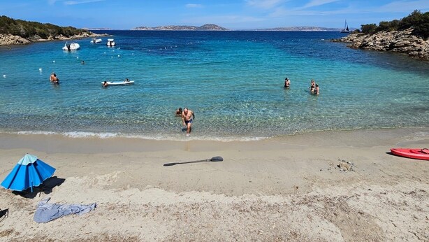 Una platja amb aigua turquesa cristal·lina, gent banyant-se i jugant a l'aigua, i un tram de sorra amb una paraigua blava i una paleta.