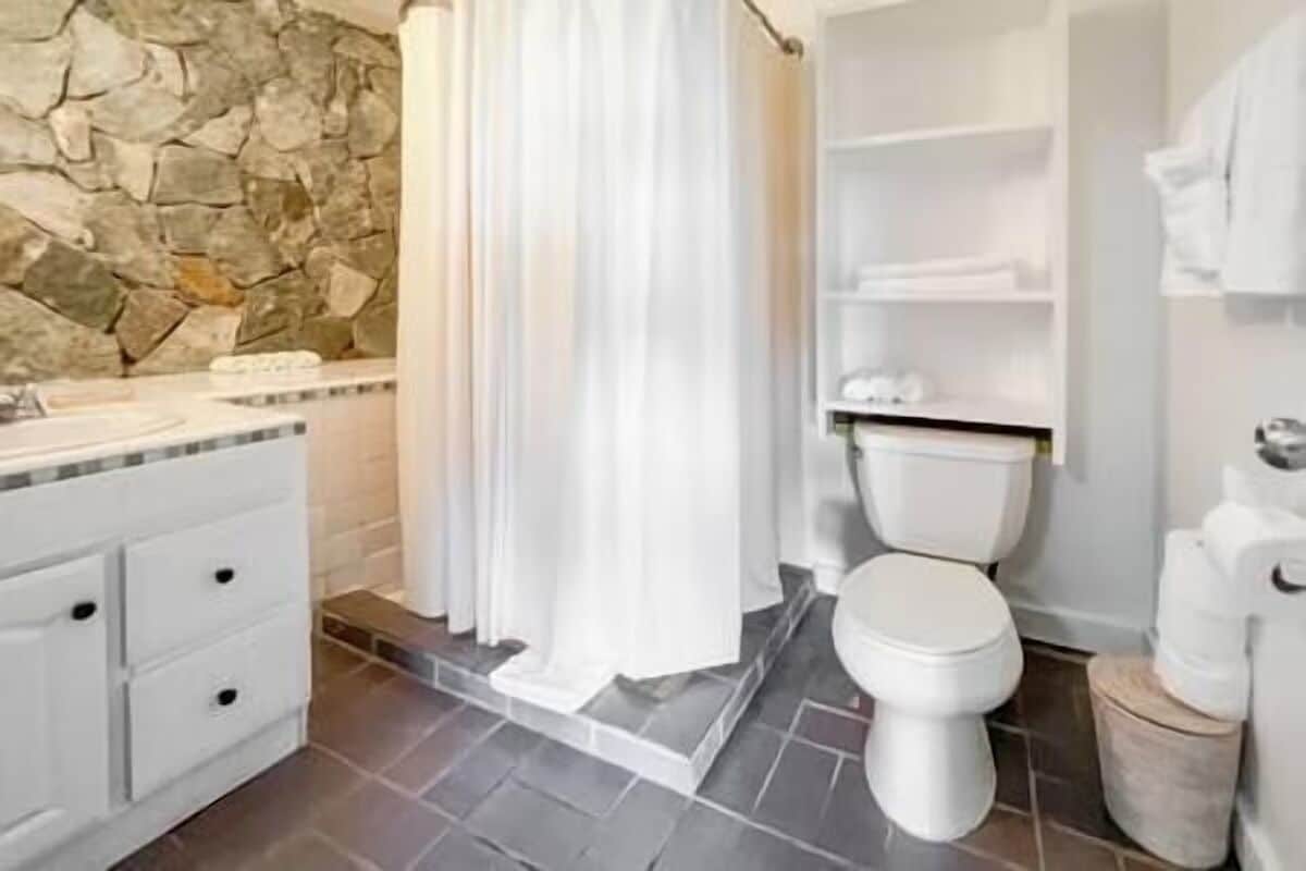 Une salle de bain avec un mur en pierre, des éléments en céramique blanche et un sol carrelé.