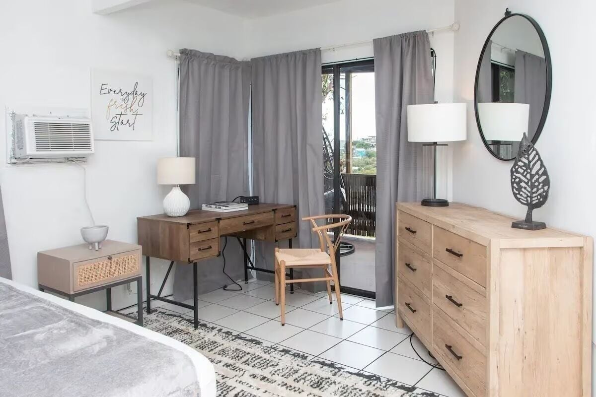 A modern bedroom with a bed, a desk, a chair, a dresser, a mirror, and a window with curtains.