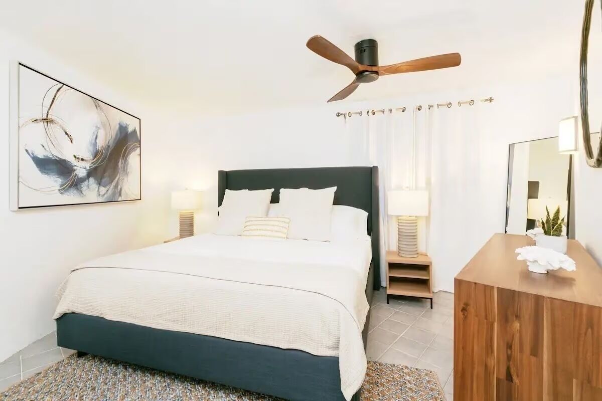Une chambre moderne avec un lit bleu foncé, du linge de lit blanc, un ventilateur de plafond, un grand tableau abstrait, une table de chevet en bois avec une lampe et un miroir.