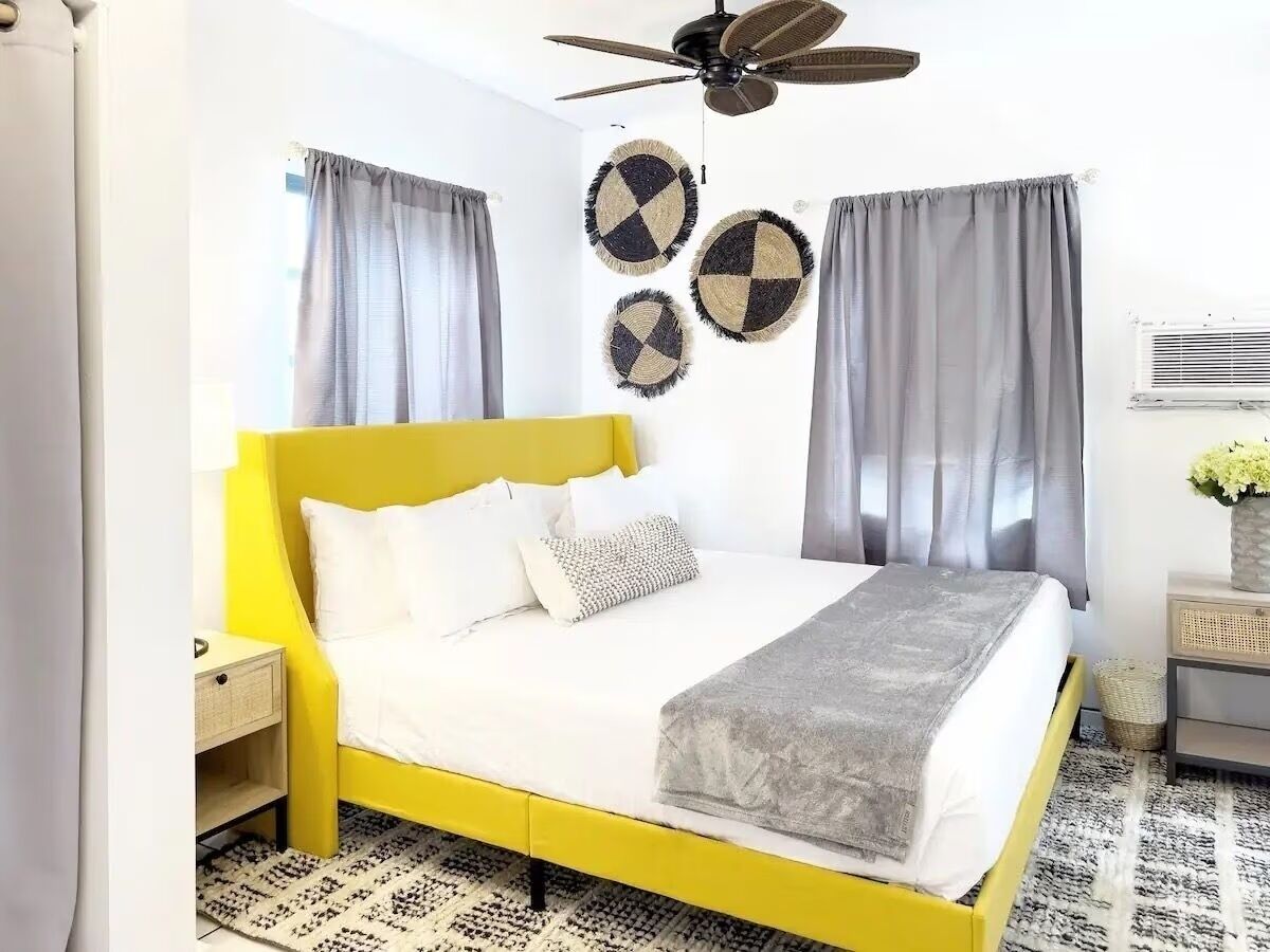 Una cama amarilla brillante con ropa de cama blanca, cortinas grises y un ventilador de techo.