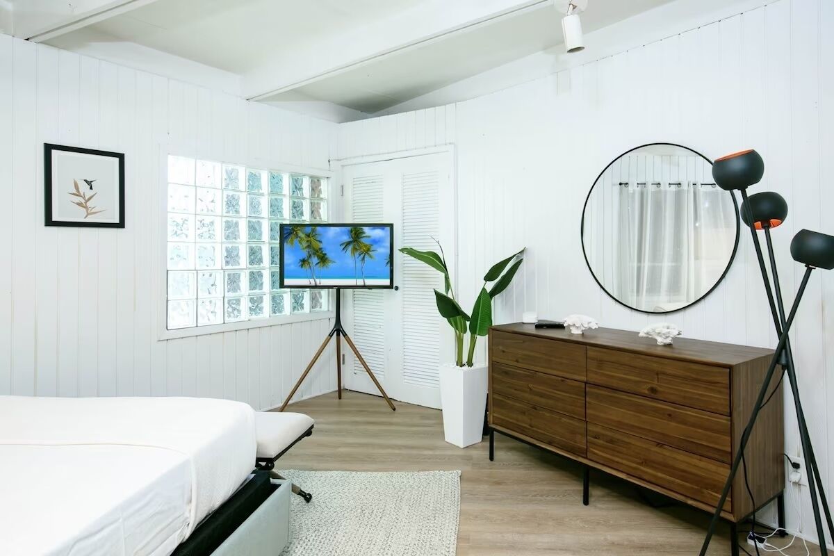 Ein Schlafzimmer mit einem Bett, einer Kommode, einem Fernseher auf einem Stativ, einem Spiegel und einem Fenster mit Jalousien.