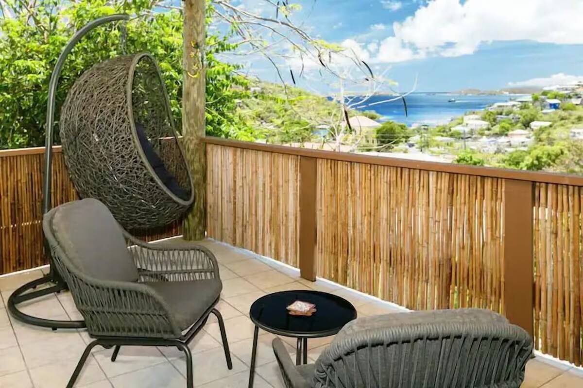 A balcony with wicker furniture, a hanging chair, and a view of the sea and buildings.