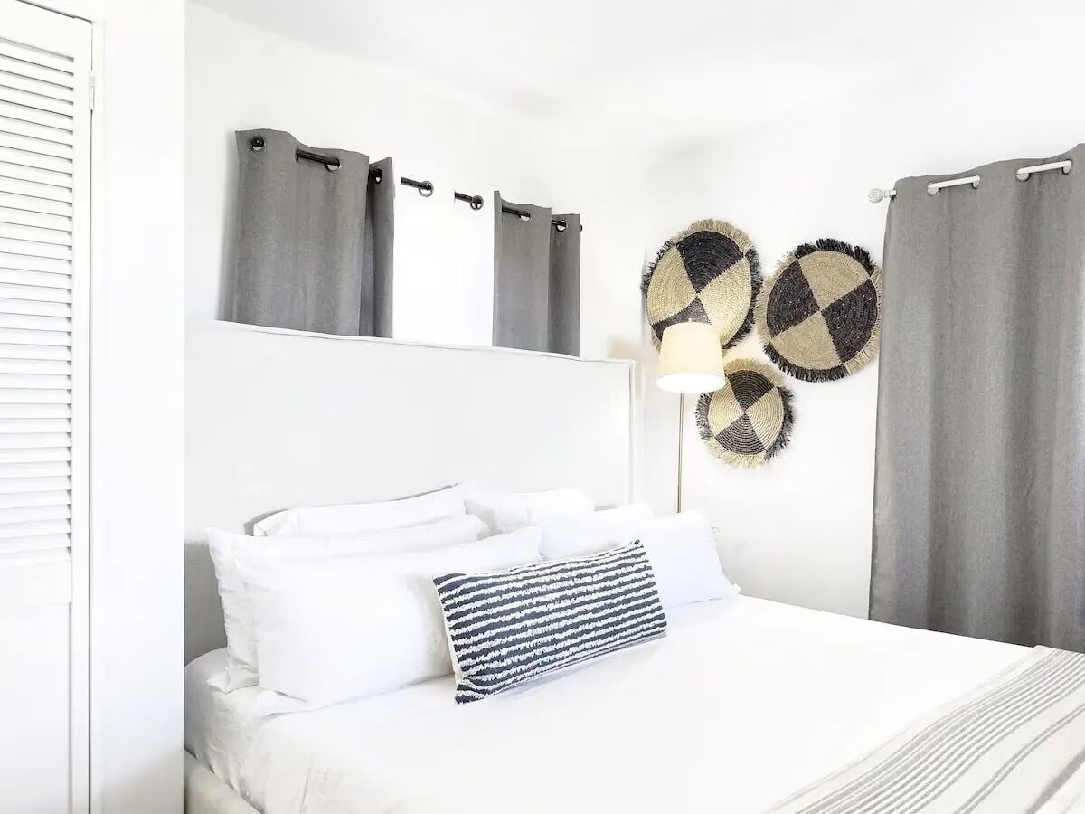 Cama bem arrumada com lençóis brancos e almofada listrada, espelho fixado na parede e duas decorações circulares.