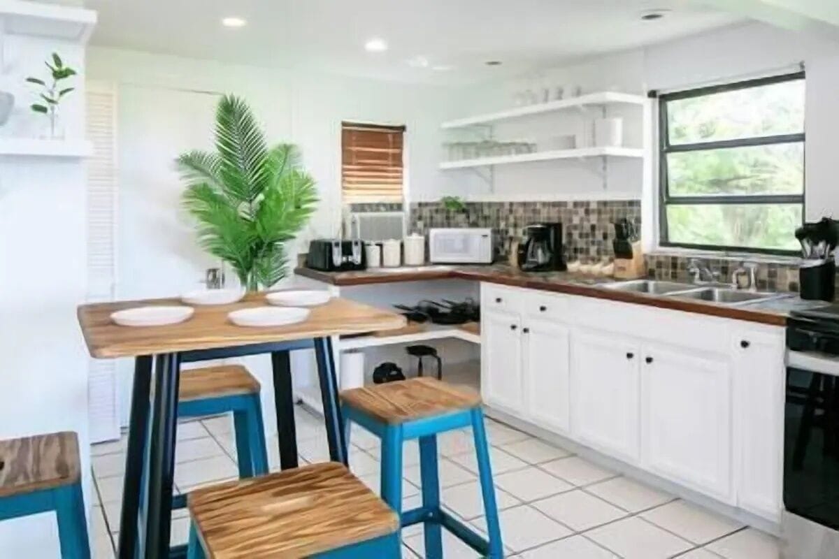 A modern kitchen with white cabinets, a wooden table, blue stools, and a large window.