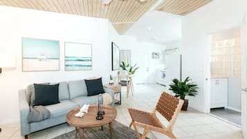 A modern living room with a sofa, coffee table, and a chair. There is a dining area with a table and chairs. The room has white walls and a wooden ceiling.