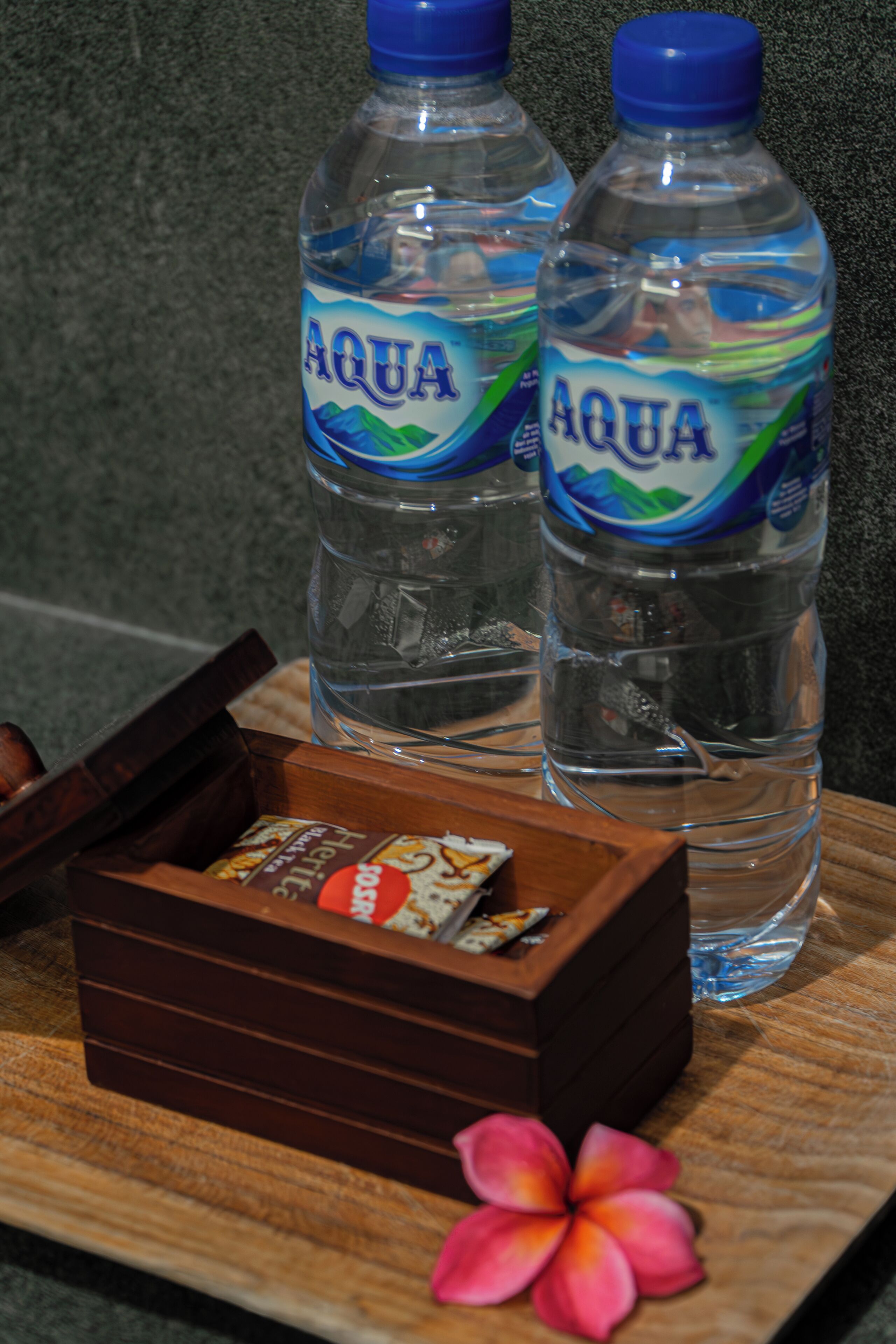 Duas garrafas de água Aqua, uma caixa de madeira com lanches e uma flor sobre uma superfície de madeira.