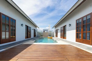 Una piscina con plataforma de madera, rodeada por muros blancos con ventanas de madera, y un cielo despejado.