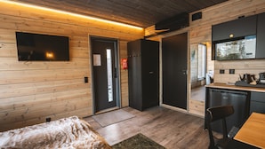 A modern, compact room with wooden walls, a flat-screen TV mounted on the wall, a small kitchenette, and a bed with a furry blanket.