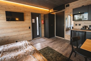 A modern, compact room with wooden walls, a flat-screen TV mounted on the wall, a small kitchenette, and a bed with a furry blanket.