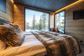 A bedroom with a large bed, wooden walls, and a view of trees through large windows. - Aurora Hill Resort (Rovaniemi)