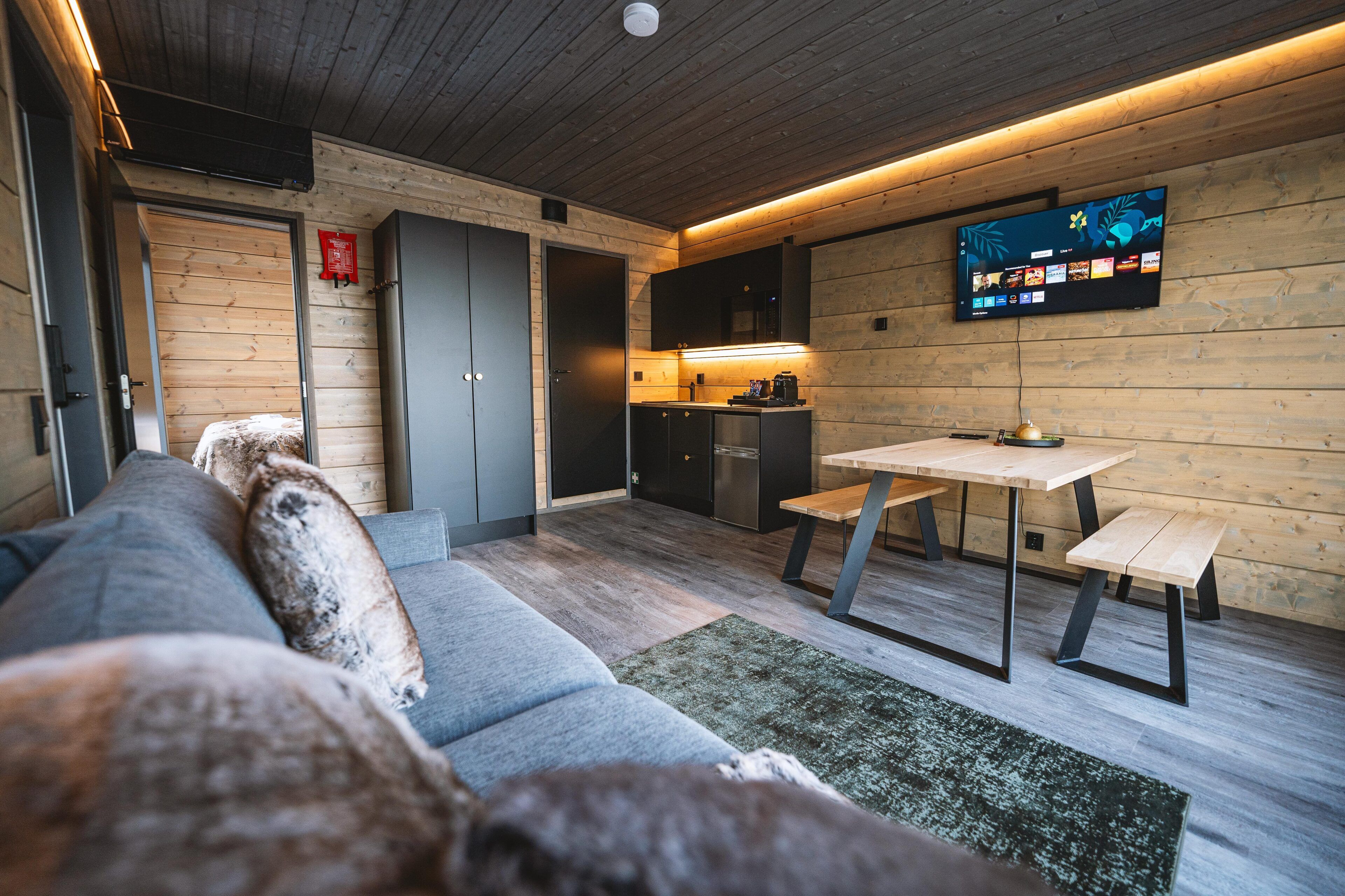 A modern kitchen with a wooden interior, a dining table with benches, and a flat-screen TV mounted on the wall.
