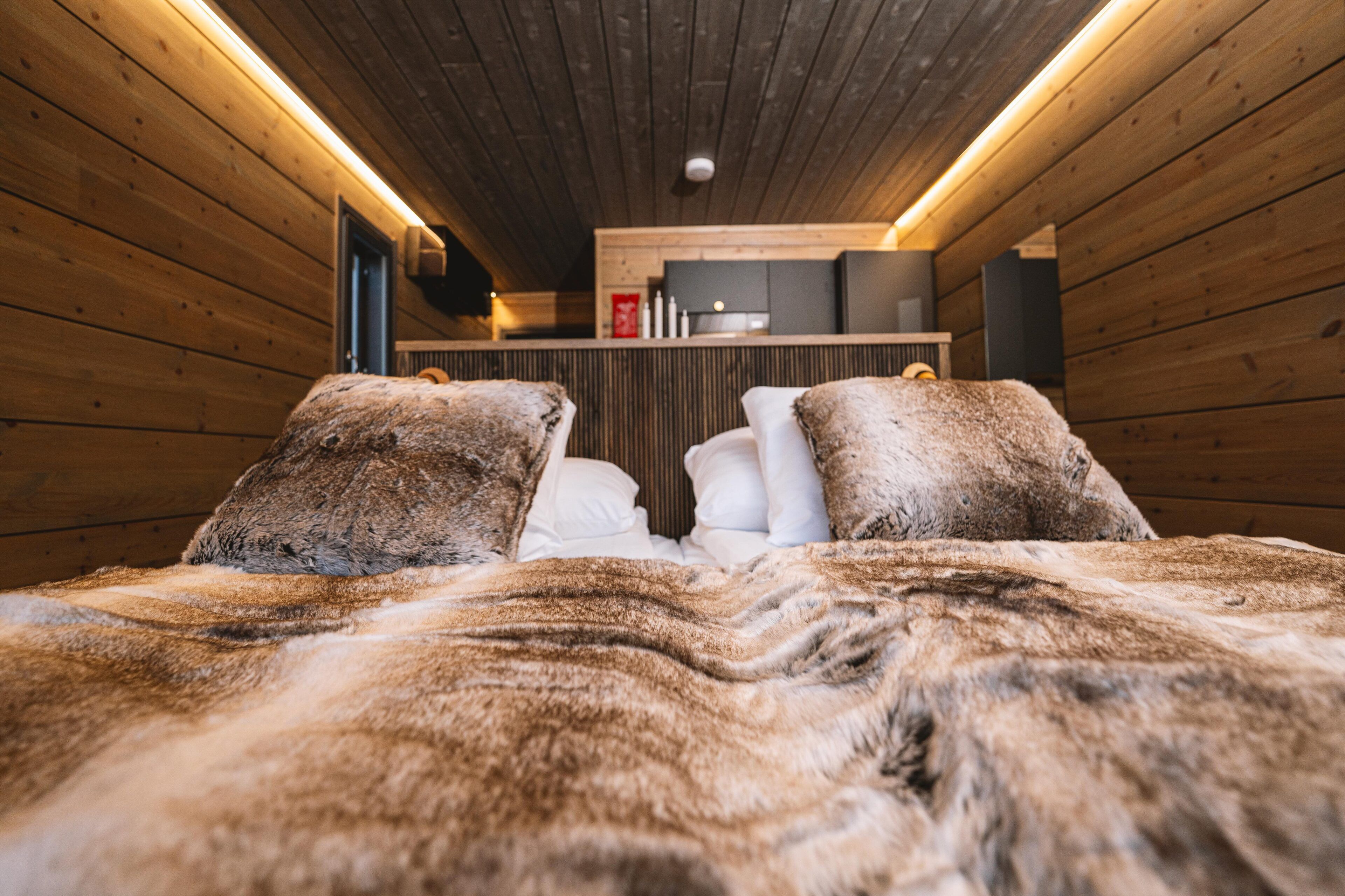 Ein gemütliches Zimmer mit Holzwänden, einem Bett mit Felldecke und einer Küchenzeile im Hintergrund.