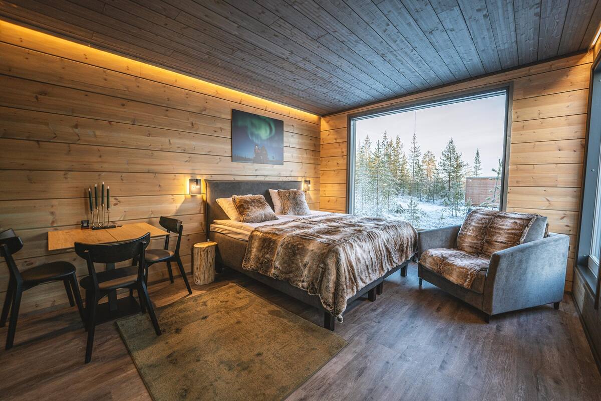 Un dormitorio acogedor con detalles de madera, un ventanal grande, una cama, un sillón y una mesita con velas.