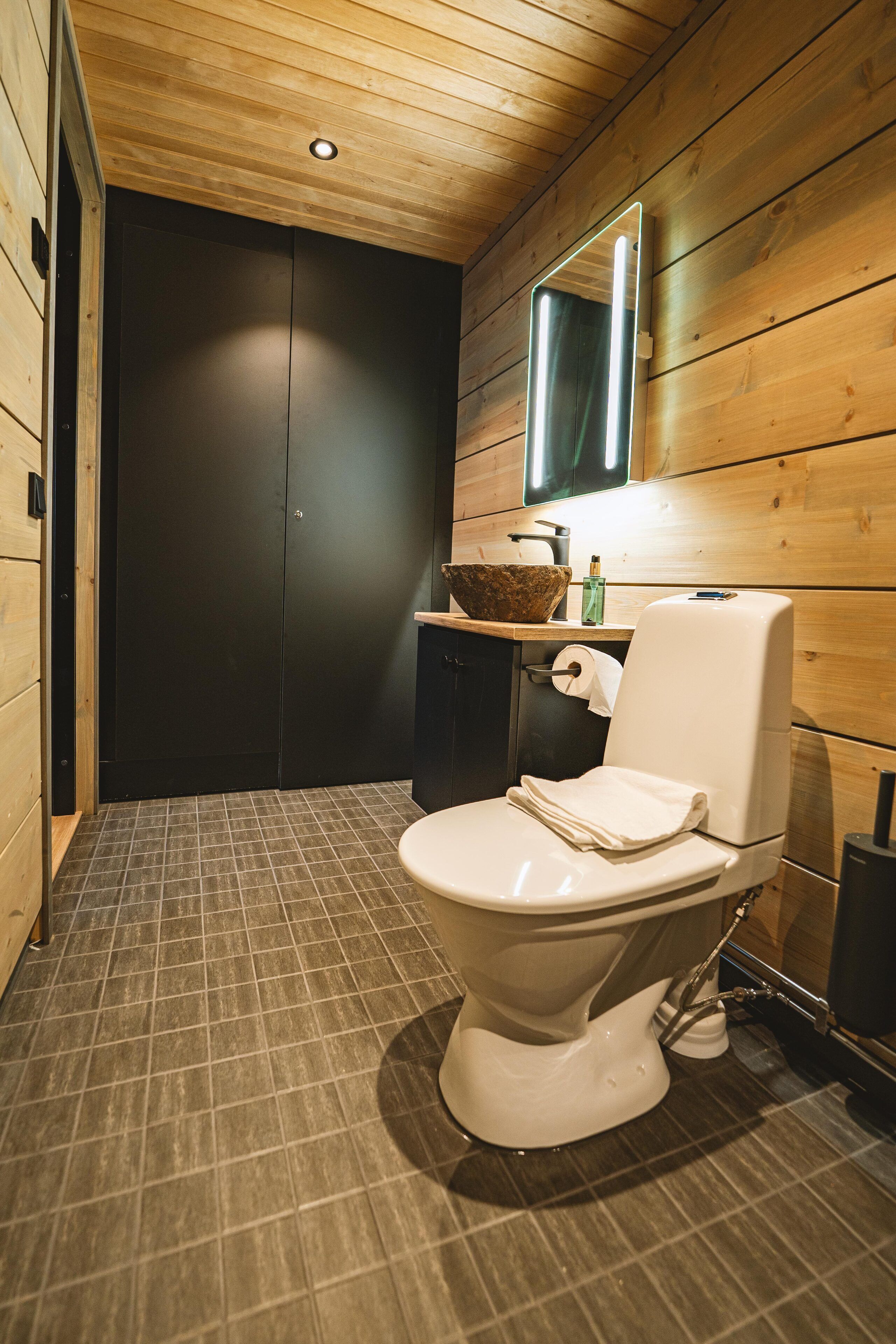 Een moderne badkamer met houten inrichting, een wit toilet, een wastafel met spiegel en een donkere deur.