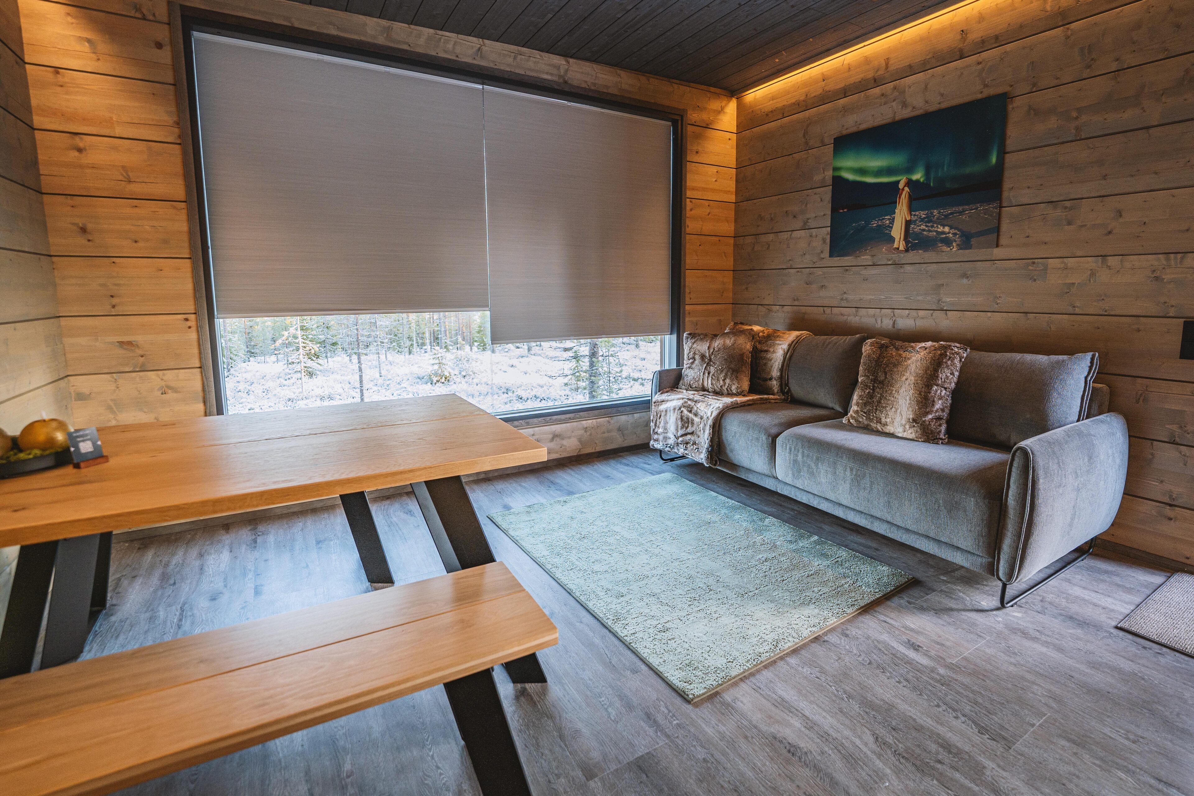 Interior acogedor de una cabaña con paredes de madera, un ventanal con vistas a un paisaje nevado, un sofá con cojines y una mesa de madera.