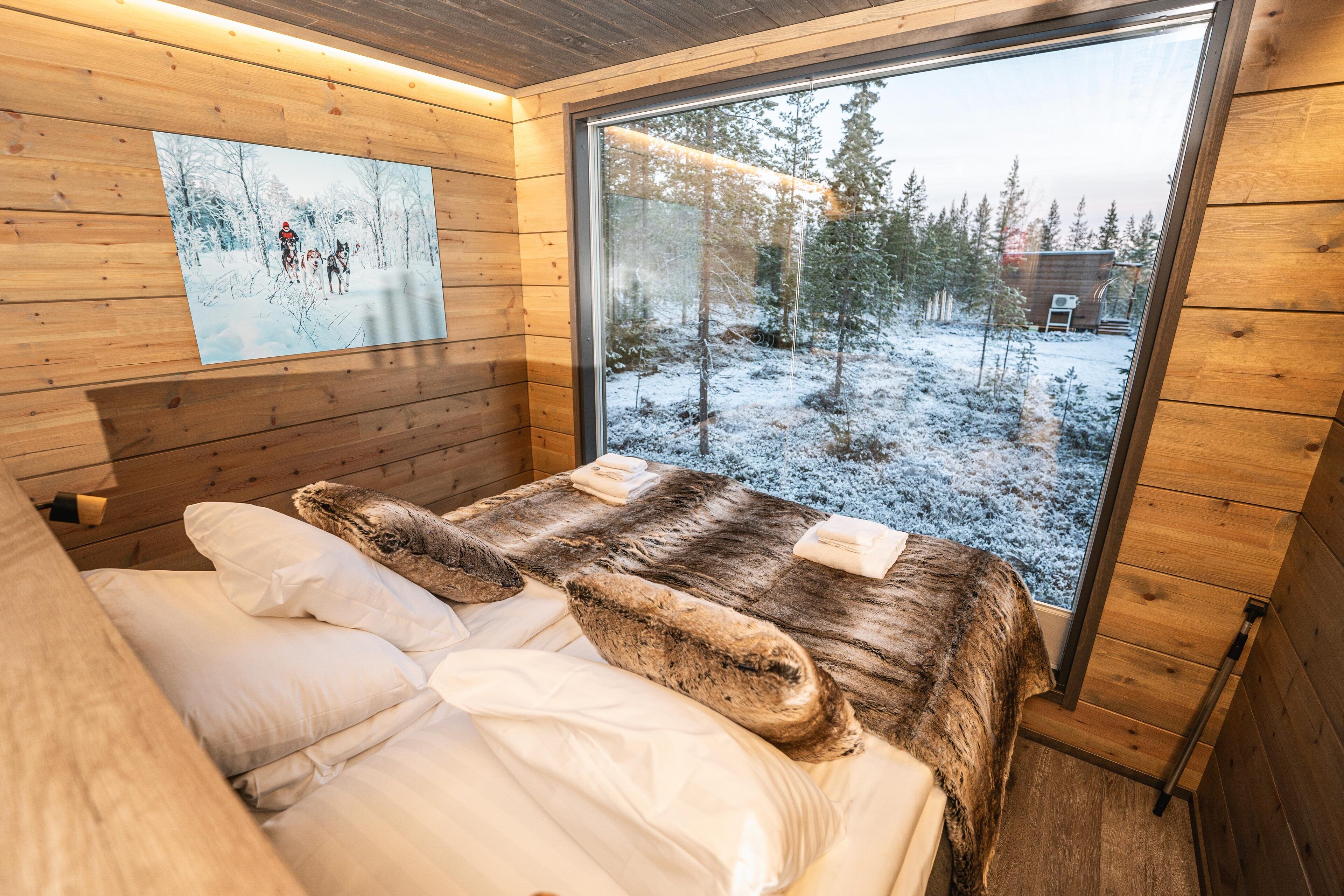 Een houten kamer met een bed, een raam en een schilderij van een besneeuwd bos.