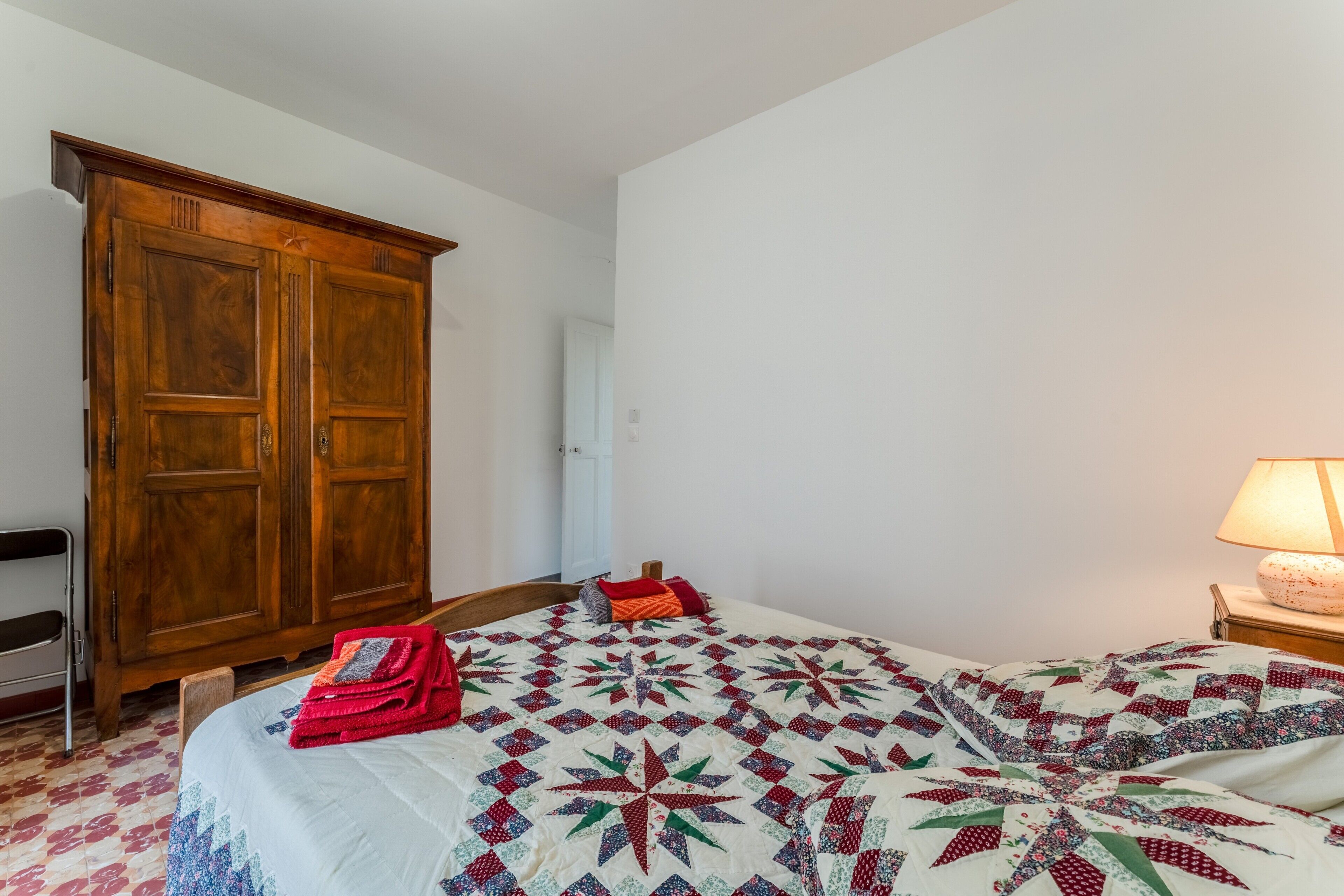 A bedroom with a bed, a wooden wardrobe, a chair, and a nightstand with a lamp.