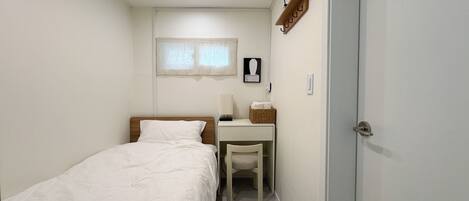 Una habitación pequeña y limpia con una cama individual, un escritorio y una silla. Hay una ventana con cortinas, un aire acondicionado de pared y una puerta.
